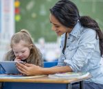 A young teacher shows her student something on a tablet at her classroom desk. Her student is elementary aged and has Down syndrome. She shows interest in the tablet.